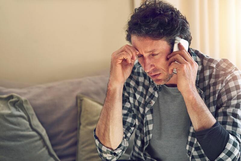 Stressed man sitting on couch holding his forehead while talking on the phone in a sunlit living room.