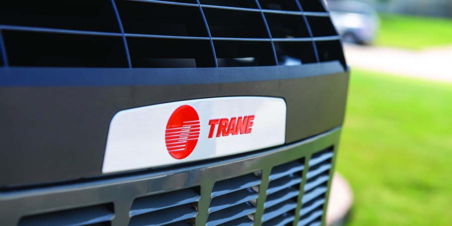 Close-up of a Trane air conditioning unit showing the brand logo, with a grassy yard and driveway in the background.