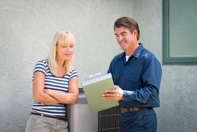 HVAC technician reviewing a clipboard with a woman next to an outdoor air conditioning unit by a stucco house wall.