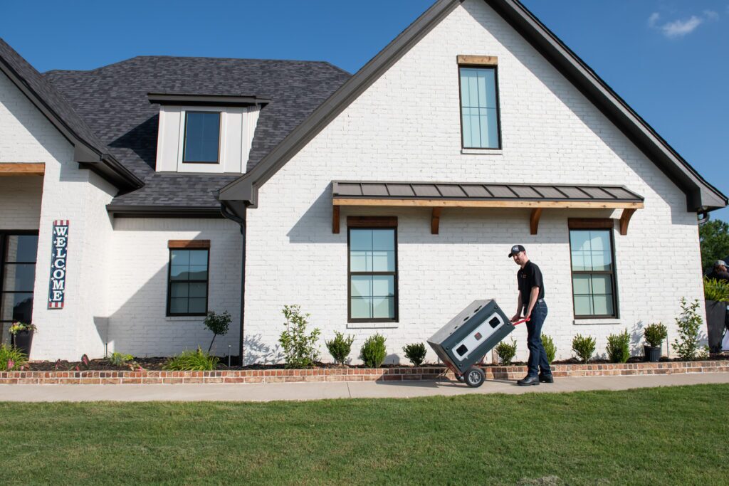 HVAC technician transporting a unit on a red hand truck along the walkway of a modern white brick home with black trim and neatly landscaped front yard on a sunny day.