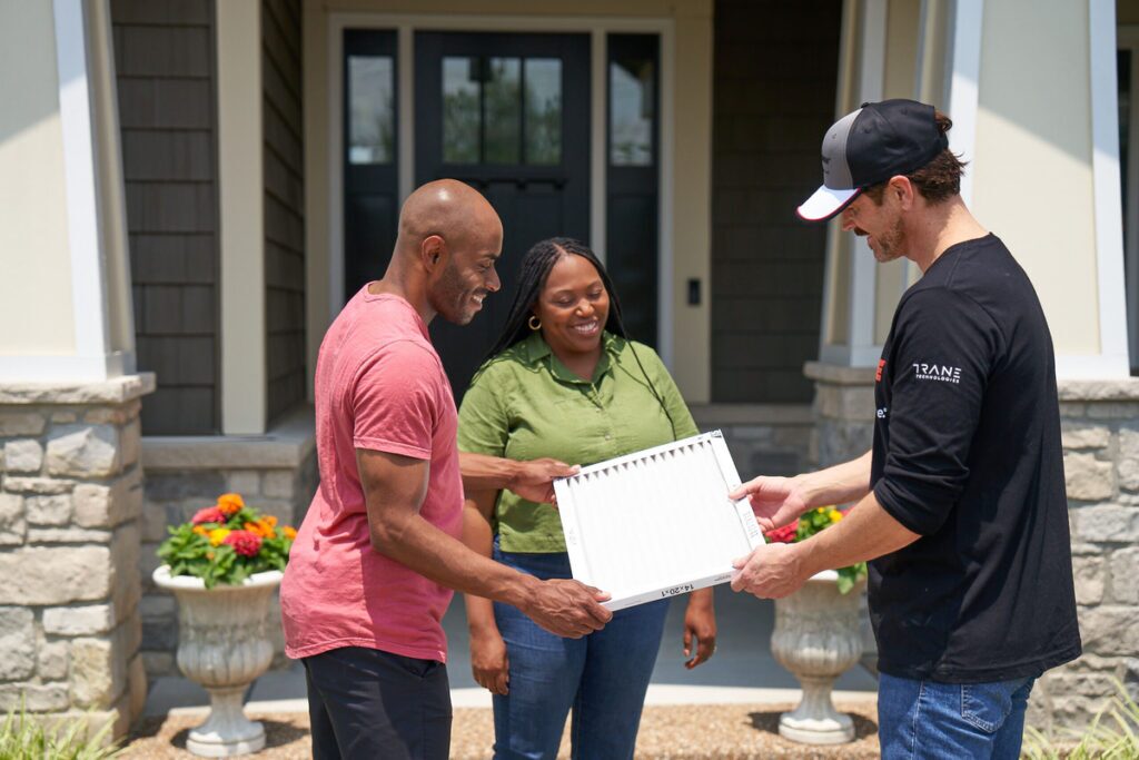 HVAC technician showing a clean air filter to a smiling couple outside their home, standing in front of a modern stone and siding entryway with flower planters on a sunny day.