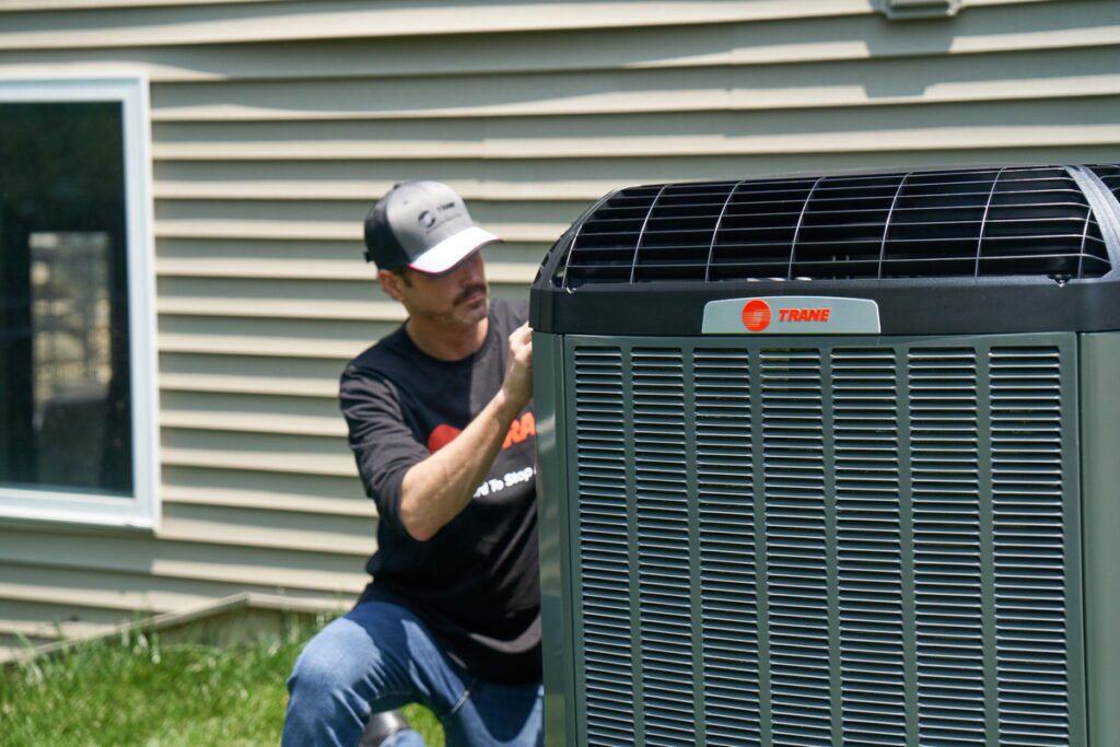 HVAC technician servicing a Trane outdoor air conditioning unit next to a residential home with beige siding on a sunny day.