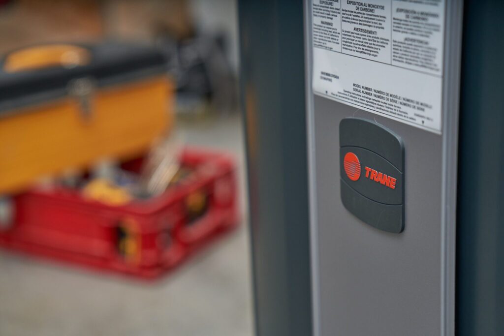 Close-up of a Trane HVAC system label on the side of a unit, with warning and model information visible above the brand logo; a toolbox and tools are blurred in the background.