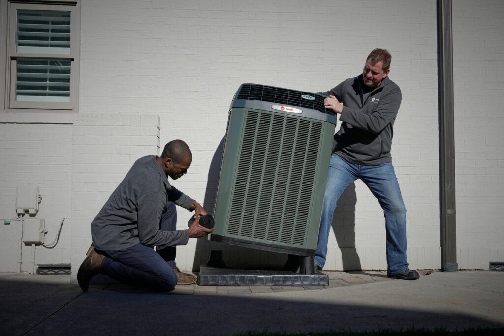 Two HVAC technicians installing or servicing a large Trane outdoor air conditioning unit next to a white brick home; one technician kneels while handling components, and the other steadies the unit.