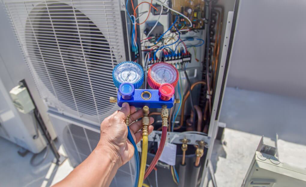 Technician holding a refrigerant manifold gauge set with red, blue, and yellow hoses connected to an open outdoor HVAC unit, showing exposed wiring and components during a service check.