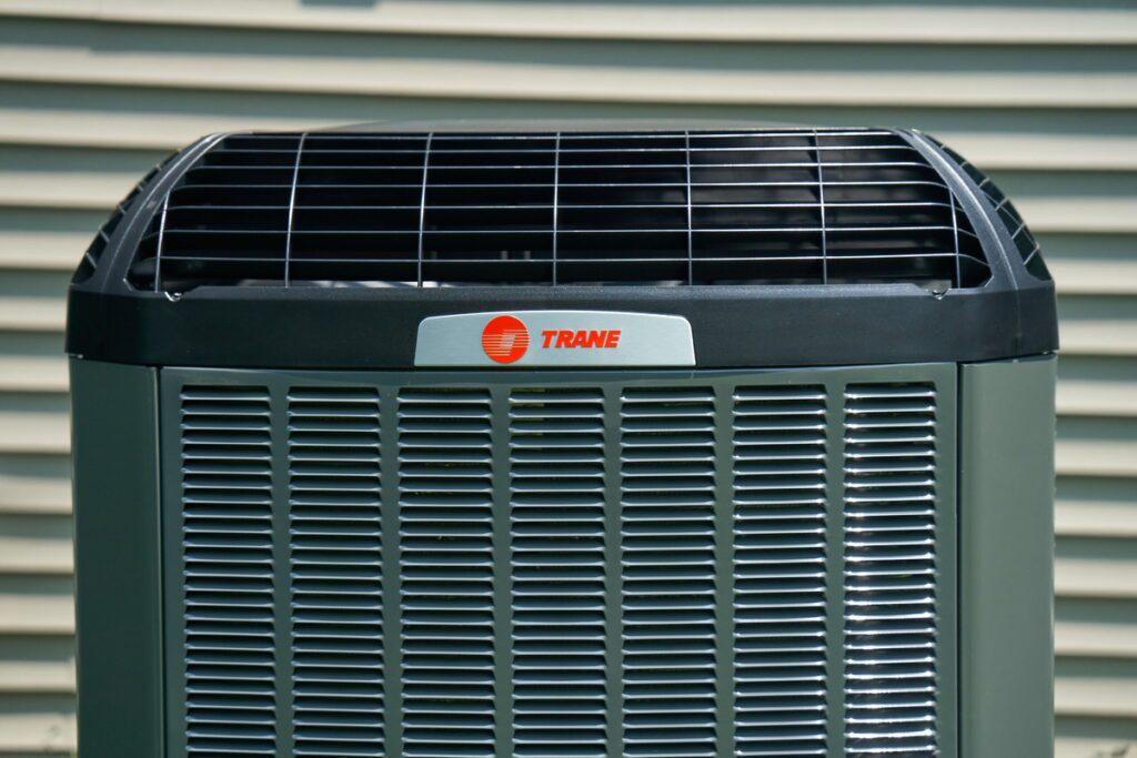 Front view of a Trane outdoor air conditioning unit placed beside a house with beige vinyl siding, featuring the brand logo and protective fan grille on top.