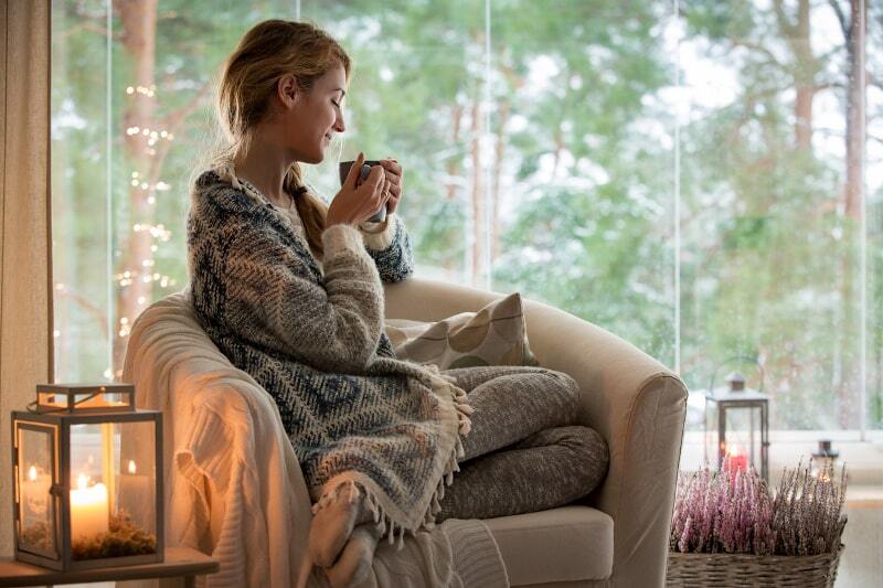 Woman relaxing on a cozy chair with a blanket and hot drink near a window, surrounded by candles and winter decor.
