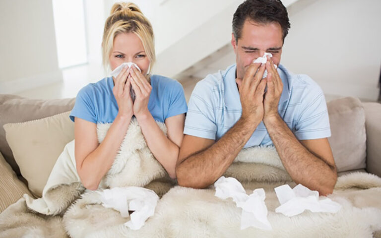 Sick couple sitting on couch under blanket, blowing noses with tissues scattered around, indicating cold or allergy symptoms.