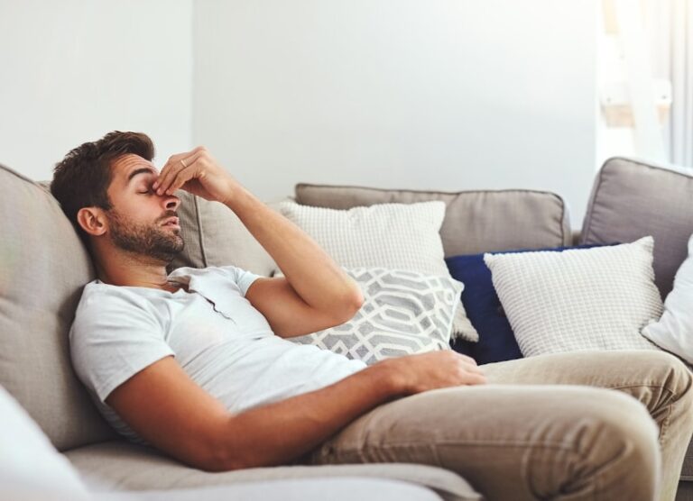 Tired man sitting on couch rubbing his eyes, surrounded by pillows in a bright living room.
