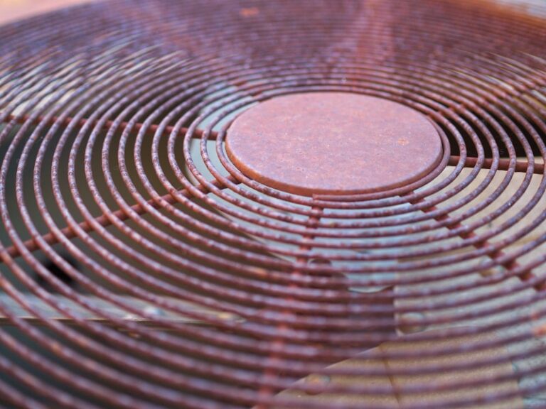 Close-up of a rusted outdoor air conditioner fan cover with circular metal grates and corroded surface.