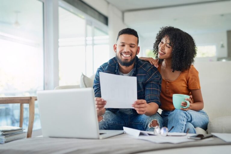 Smiling couple reviewing documents at home with laptop, coffee mug, and papers on table in bright modern living room.