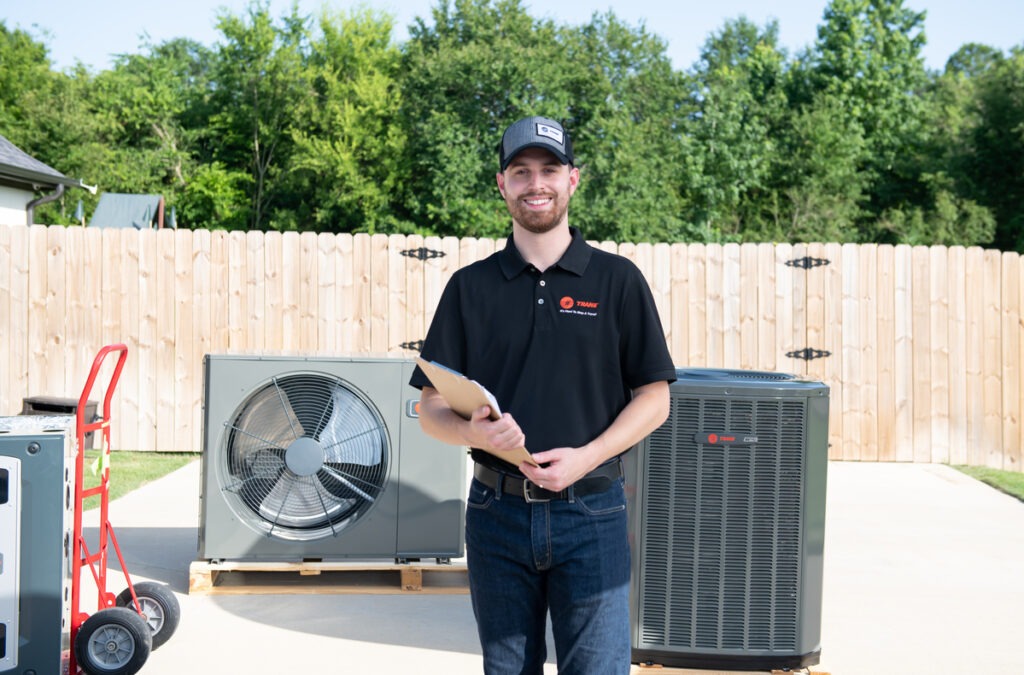 Smiling HVAC technician holding a clipboard while standing in a residential driveway, surrounded by Trane air conditioning and heat pump units, with a wooden privacy fence and trees in the background.