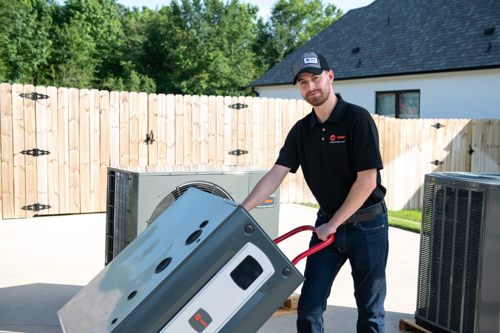 HVAC technician using a red hand truck to move a Trane unit in a residential driveway, surrounded by other HVAC equipment and a wooden privacy fence with trees in the background.