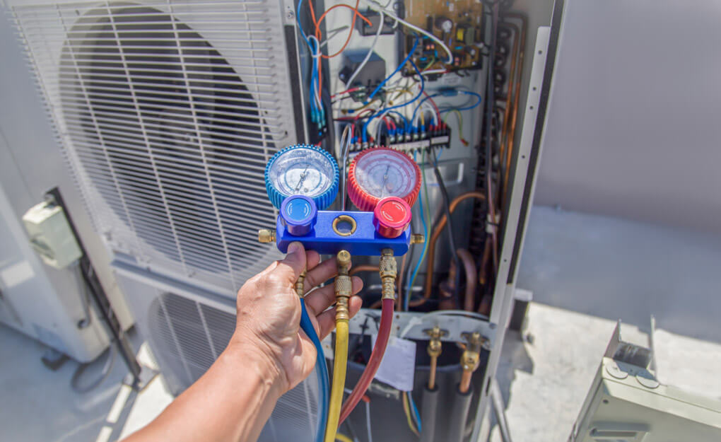 Technician holding a refrigerant manifold gauge set with red, blue, and yellow hoses connected to an open outdoor HVAC unit, showing exposed wiring and components during a service check.